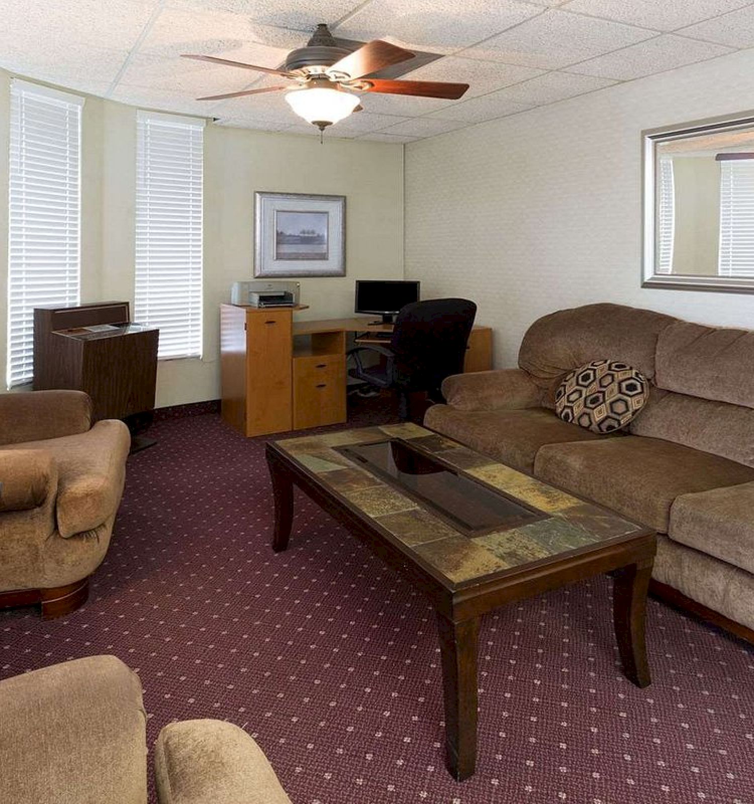 A living room with brown furniture, a coffee table, a desk with a computer, wall art, and a large mirror, ceiling fan, and carpeted floor.