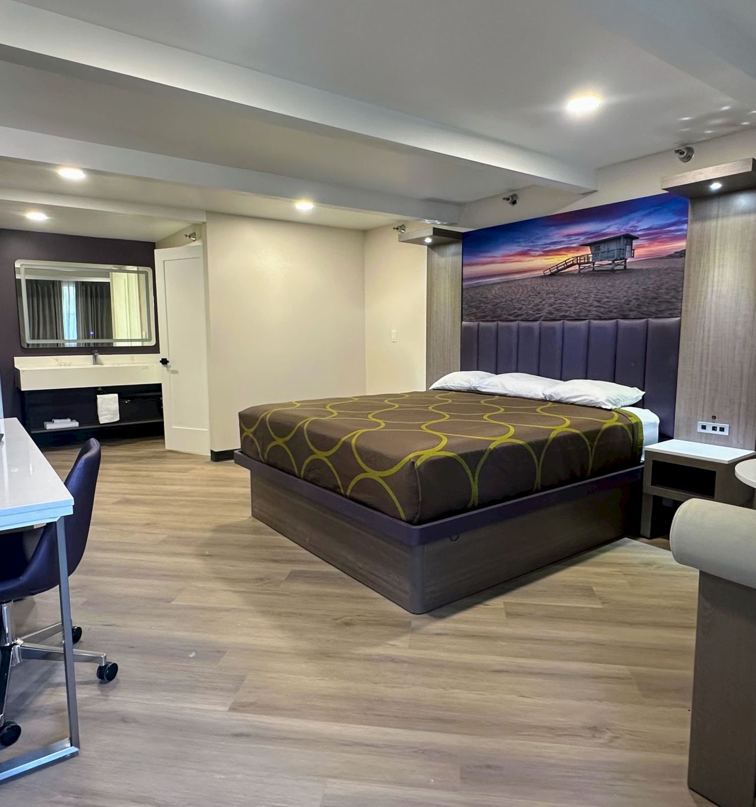A modern hotel room with a bed, desk, chair, wall-mounted TV, table, and chairs, with a scenic photograph above the bed.