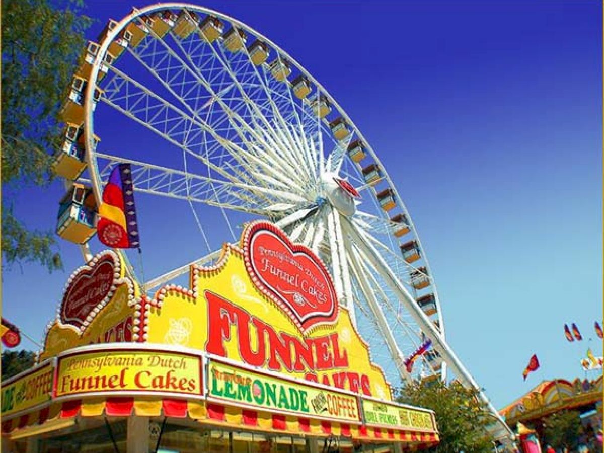 The image shows a large Ferris wheel at a fair, with a brightly colored stand offering funnel cakes and lemonade in the foreground.