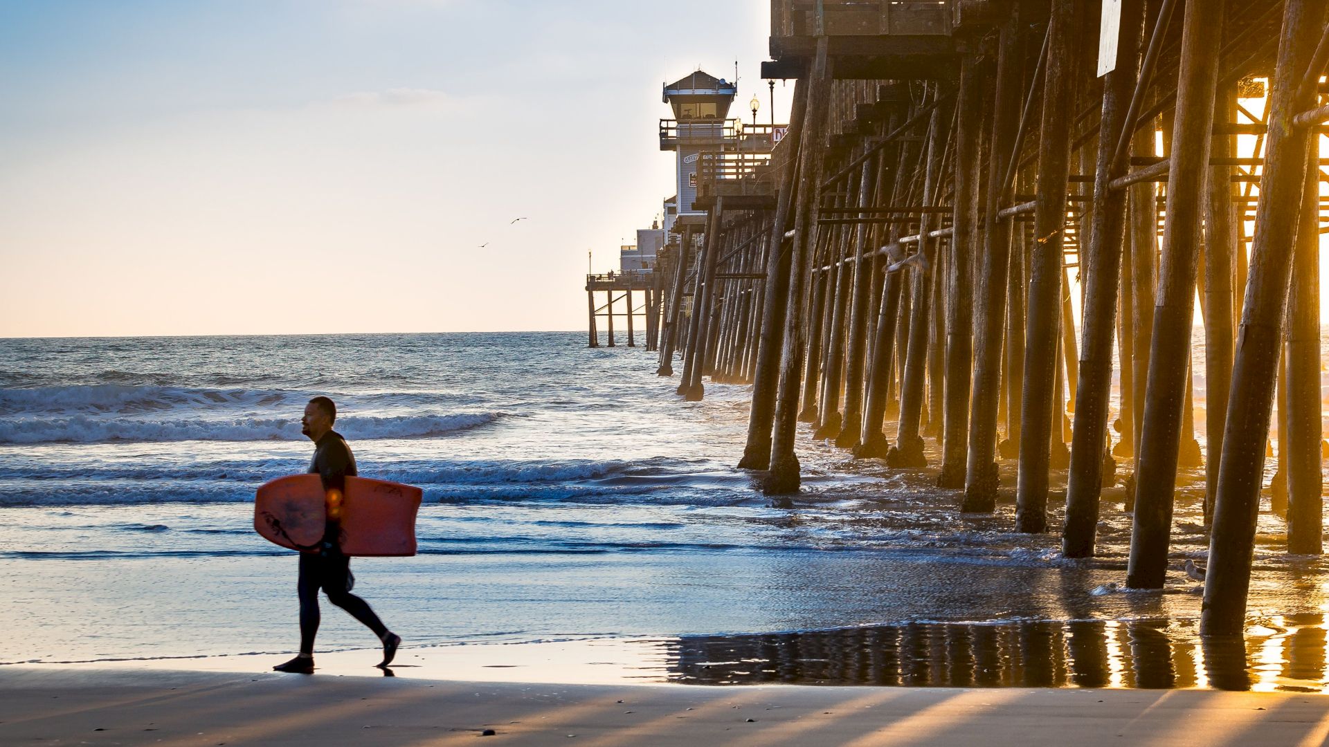 A person carrying a surfboard walks along the beach near a pier during sunset.