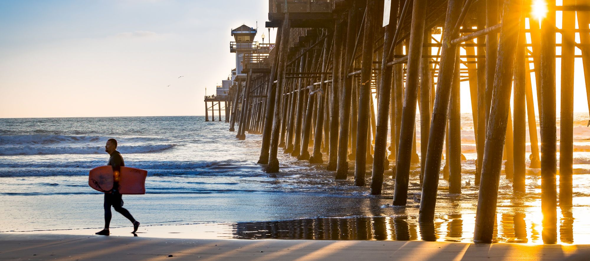 A person carrying a surfboard walks along the beach near a pier during sunset.