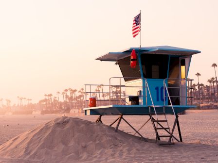 A lifeguard tower on a sandy beach with an American flag on top and palm trees in the background, at sunrise or sunset.