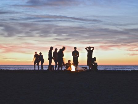 A group of people is gathered around a campfire on a beach at sunset, with the ocean and colorful sky in the background.