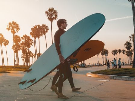 Two surfers carrying surfboards walk along a palm tree-lined path at sunset.