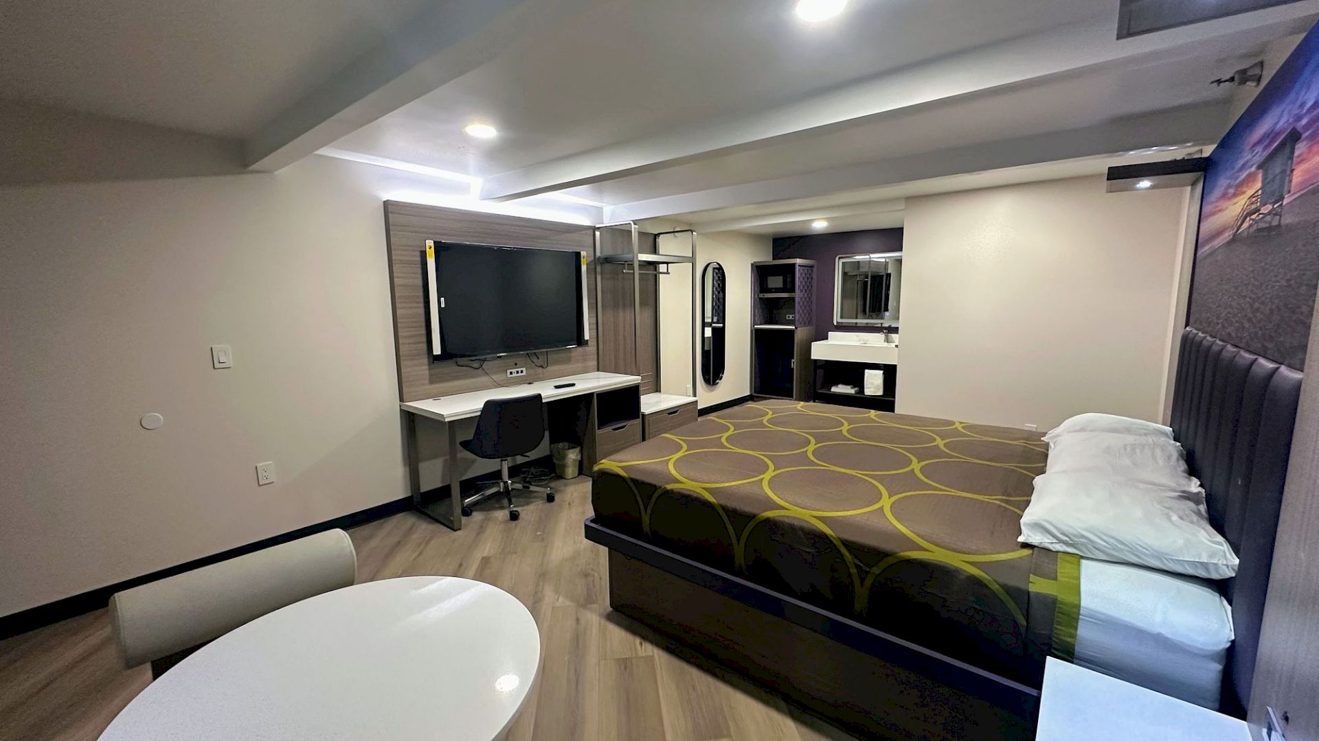 The image shows a modern hotel room with a bed, desk, chair, TV, and a small table. Neutral colors and streamlined decor create a cozy atmosphere.