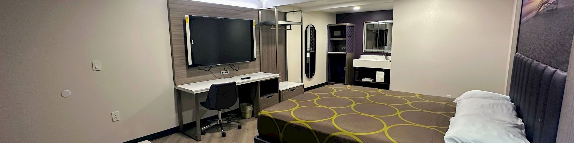 The image shows a modern hotel room with a bed, desk, chair, TV, and a small table. Neutral colors and streamlined decor create a cozy atmosphere.