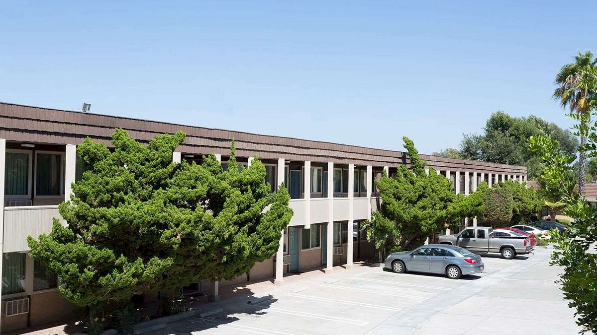 The image shows a two-story building with trees in front and cars parked in a lot. The sky is clear and sunny, suggesting a warm day.