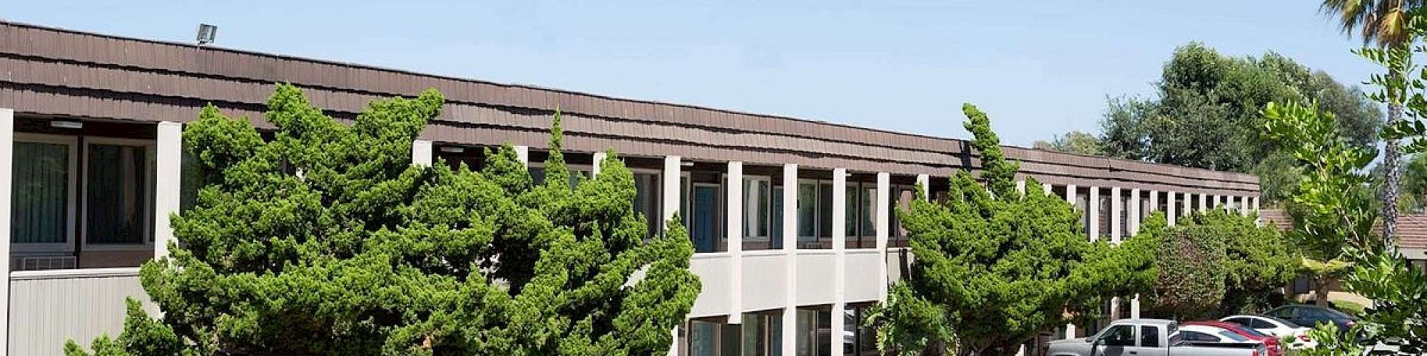 The image shows a two-story building with trees in front and cars parked in a lot. The sky is clear and sunny, suggesting a warm day.