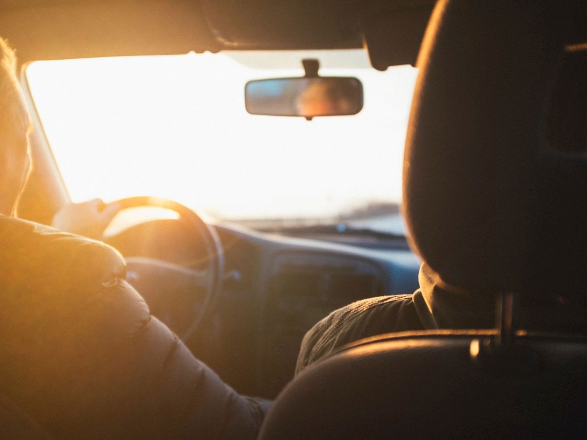 The image shows two people in the front seats of a car, with the driver steering and a bright sunset visible through the windshield.