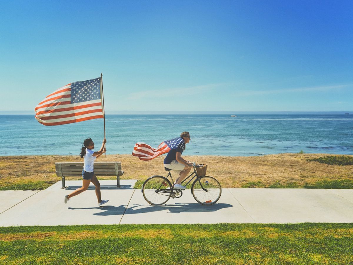 Two people are by the seaside, one is riding a bicycle with an American flag cape, and another is running with an American flag.