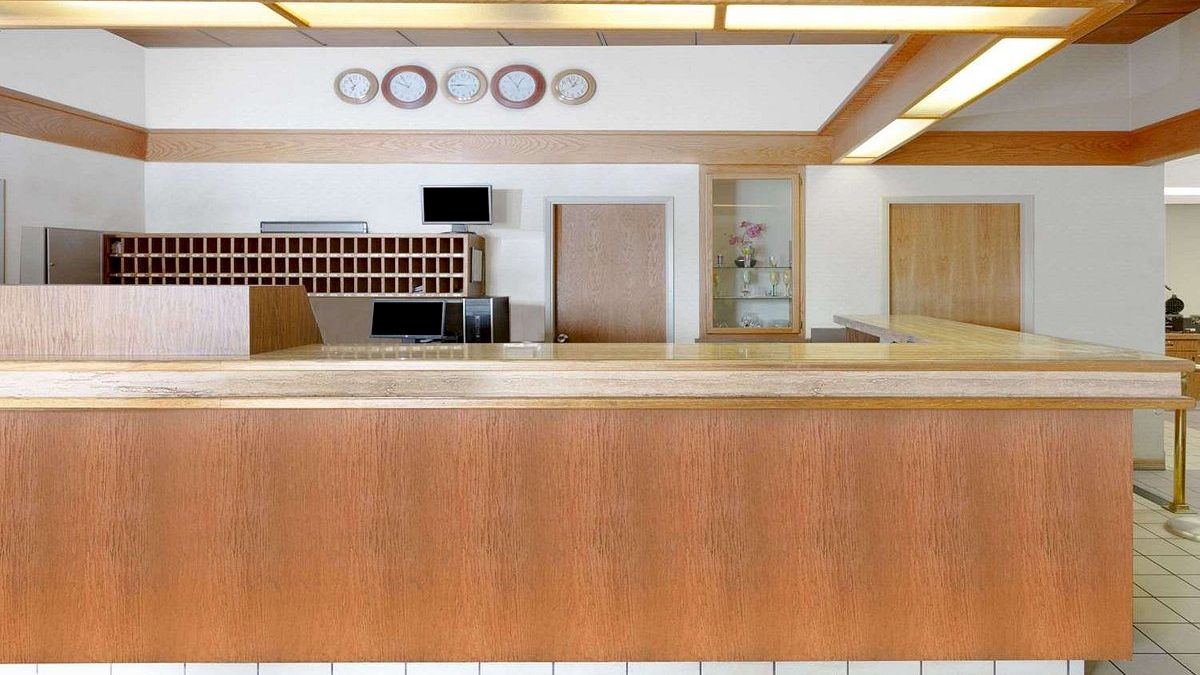 This image shows a hotel reception desk with wooden accents, a check-in counter, a key holder board, clocks on the wall, and tiled flooring.