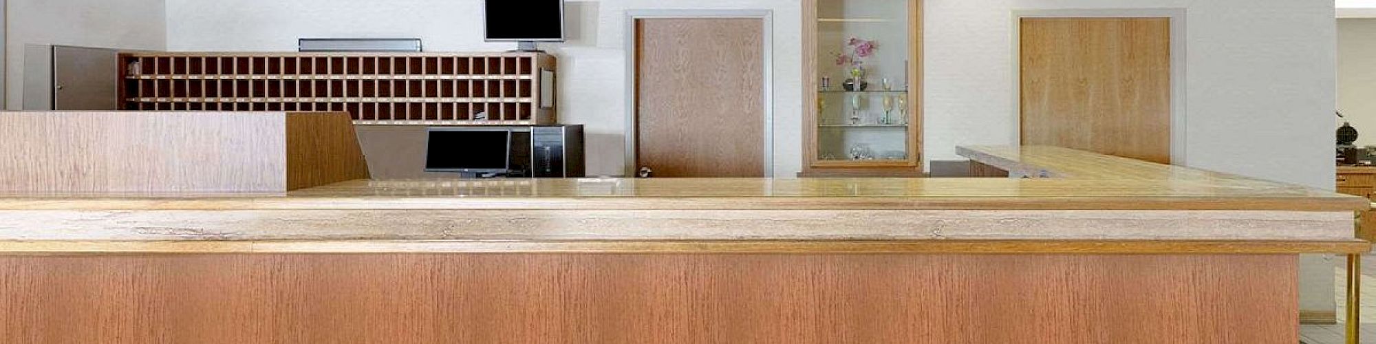 This image shows a hotel reception desk with wooden accents, a check-in counter, a key holder board, clocks on the wall, and tiled flooring.