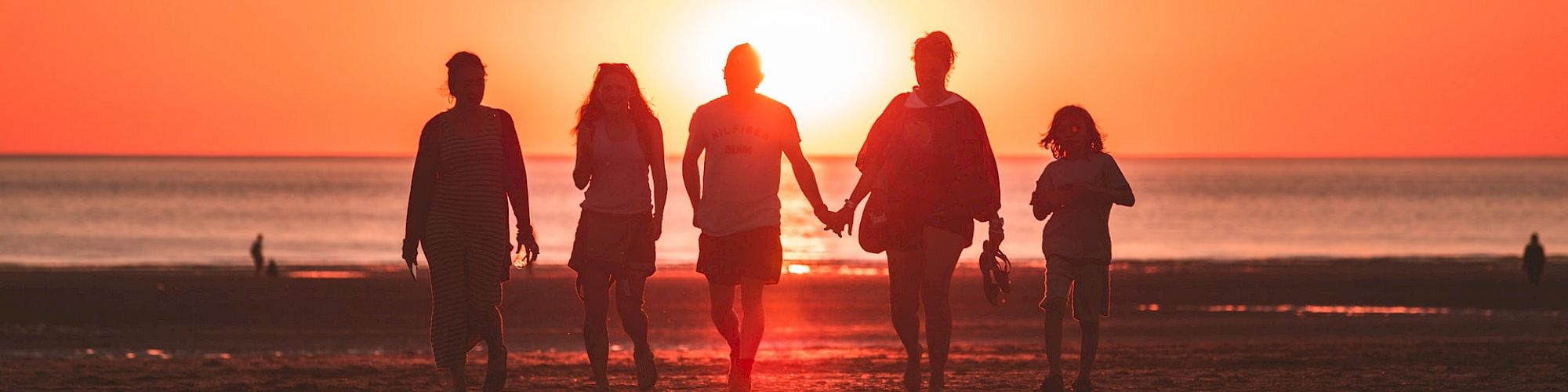 A group of five people, silhouetted against a vibrant sunset, walk along a sandy beach, hand in hand, with a calm ocean in the background.