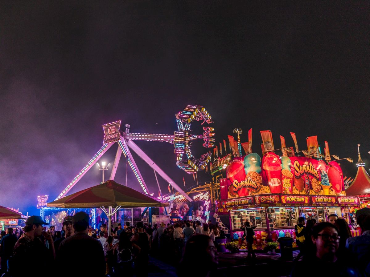 A vibrant carnival scene at night with colorful rides and games, including a Ferris wheel and stalls, surrounded by a crowd of people enjoying.