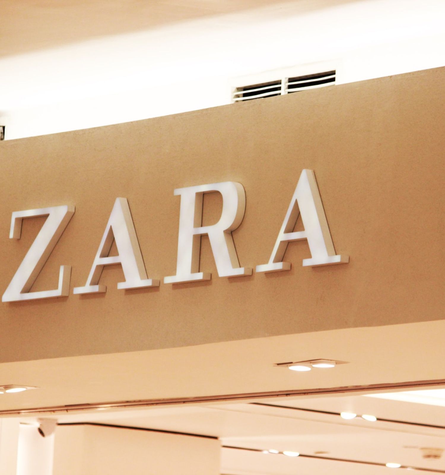 The image shows the sign for a Zara store, prominently displayed in white letters on a beige wall inside a shopping mall.