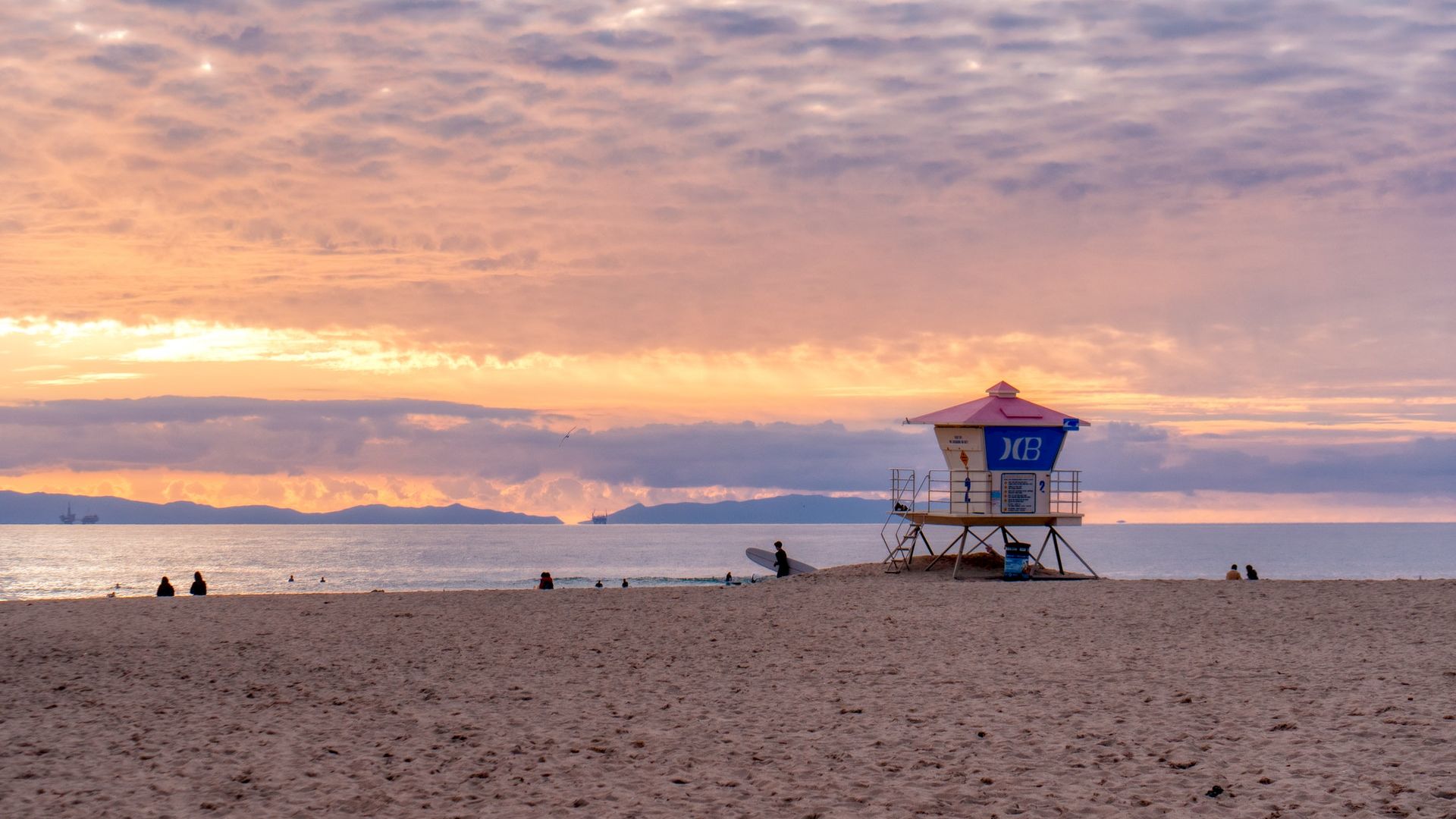 A lifeguard tower stands on a sandy beach at sunset, with a dramatic sky and calm ocean in the background, and a few people scattered on the shore.
