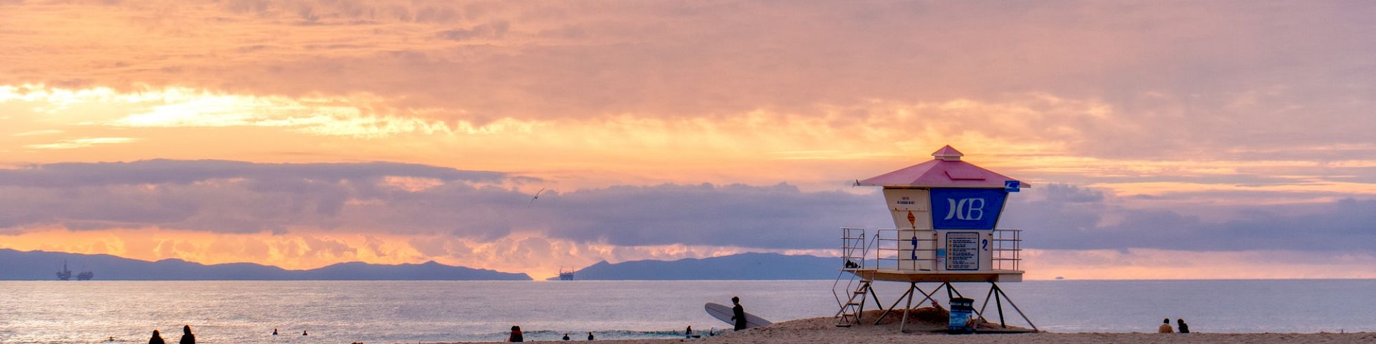 A lifeguard tower stands on a sandy beach at sunset, with a dramatic sky and calm ocean in the background, and a few people scattered on the shore.