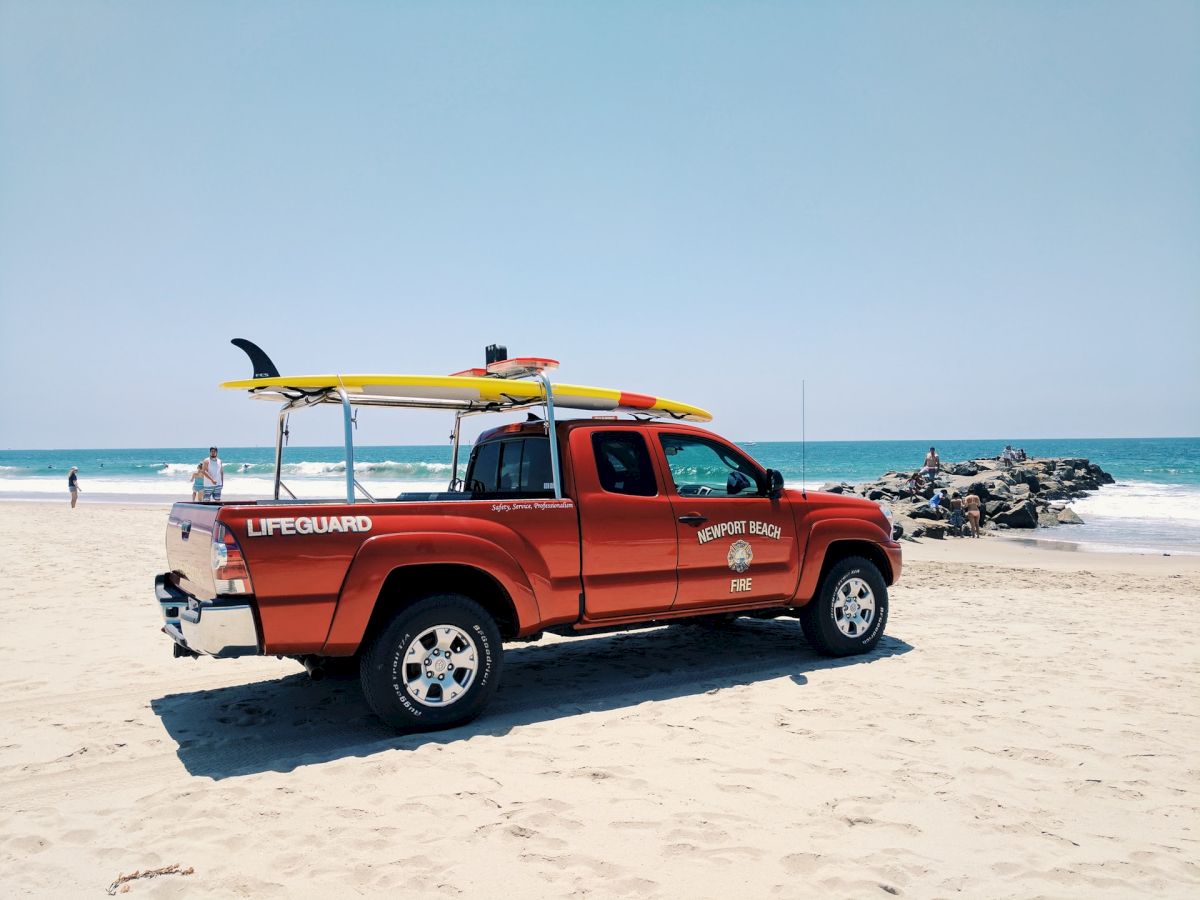 A red lifeguard truck is parked on a beach near the ocean, equipped with surfboards on the roof.