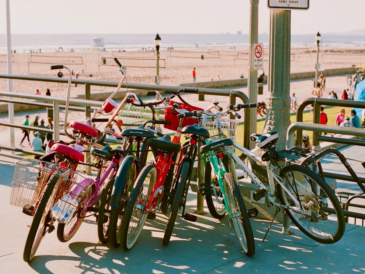 A row of colorful bicycles is parked near a beach setting with people in the background and a boardwalk visible, overlooking the ocean.