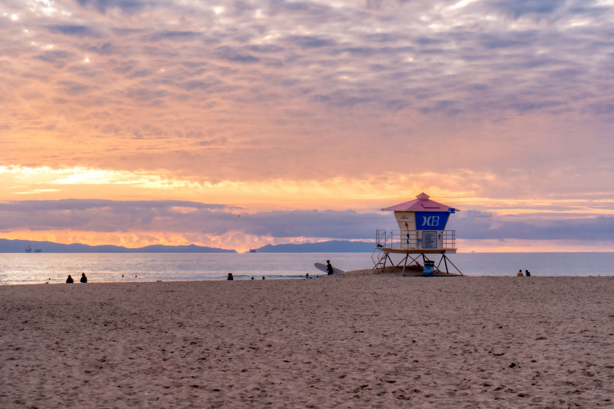 A lifeguard tower stands on a sandy beach at sunset with a cloudy sky while people sit near the shoreline and distant mountains are visible.