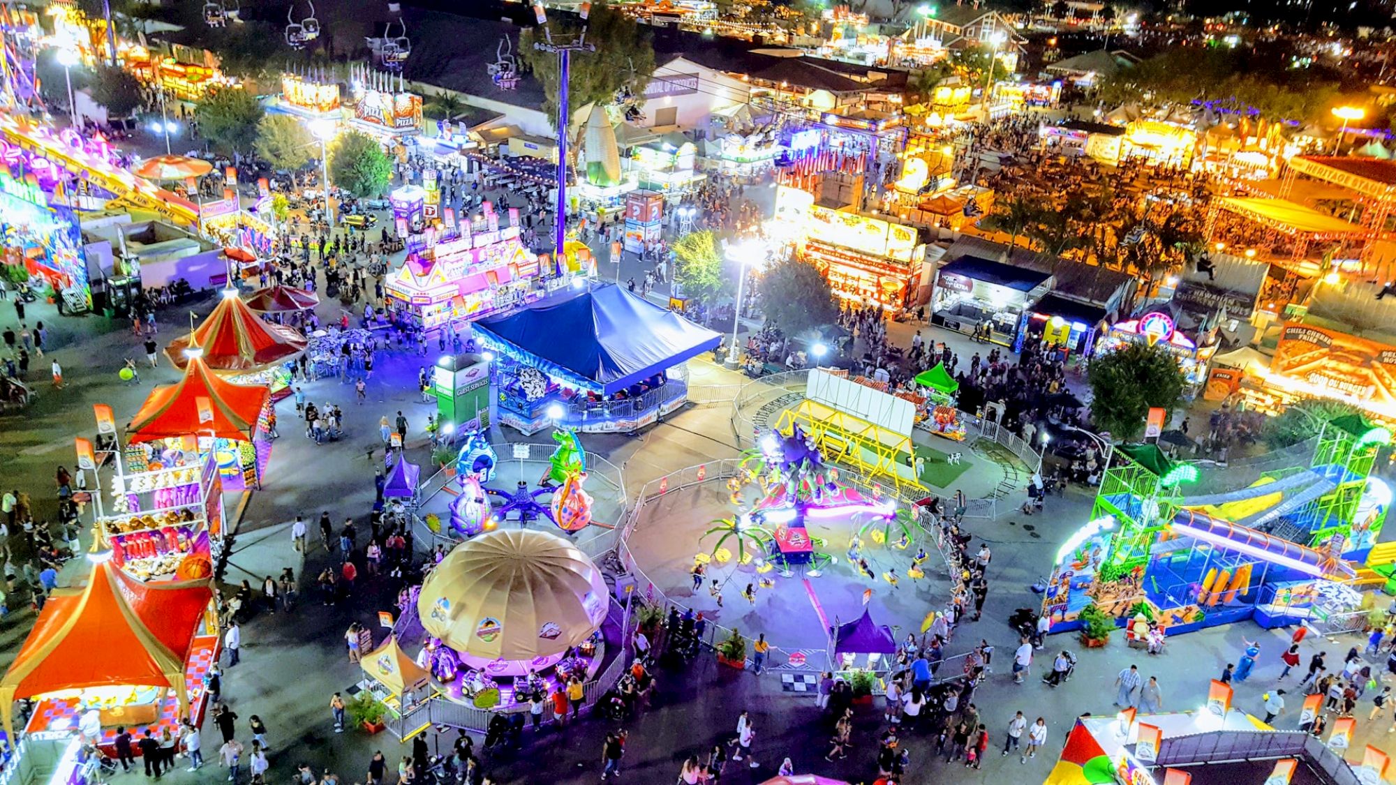 The image shows a vibrant nighttime carnival scene with colorful tents, rides, and numerous people enjoying the festivities.