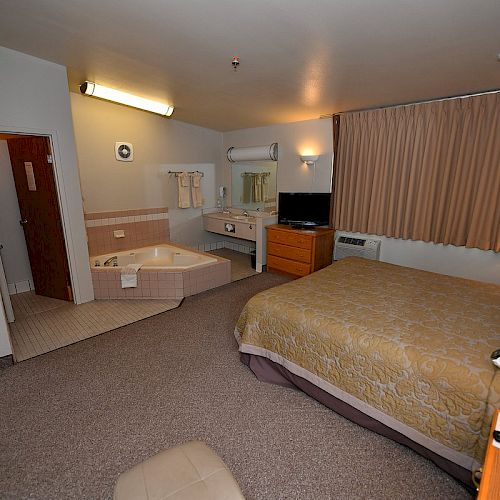 The image shows a hotel room with a bed, a jacuzzi tub, a TV on a dresser, a vanity area, and a window with curtains.