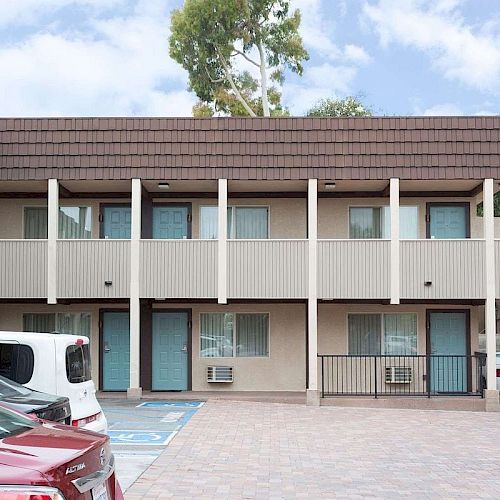 A two-story motel with parking spaces, including accessible parking, in front of the building, with trees and a blue sky in the background.