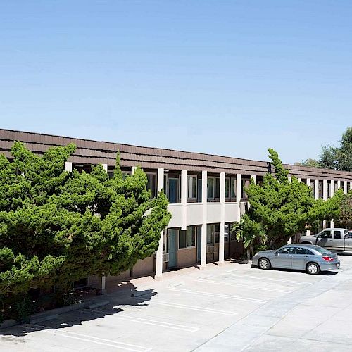 A two-story building is surrounded by trees, with a parking lot in front, containing a few cars under a clear blue sky.