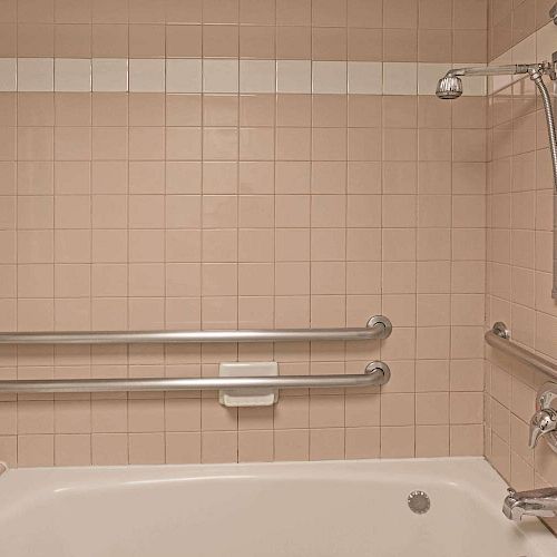 A bathroom with pink square tiles, a bathtub, and a handheld shower. There are two grab bars and a soap holder on the wall.