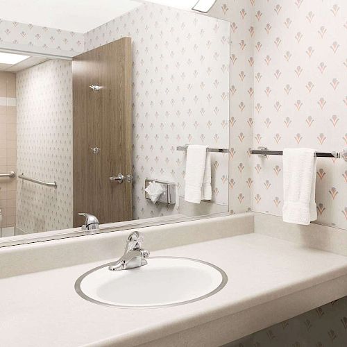 This image depicts a clean bathroom with a sink, mirror, towels, and a door leading to a toilet. The walls have a patterned wallpaper.