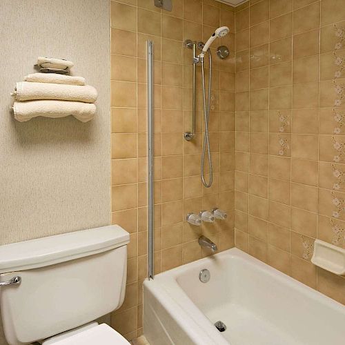 This image shows a bathroom with tan tiles, a bathtub-shower combo, a toilet, and folded towels on a shelf above the toilet.