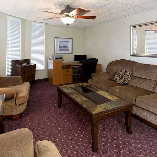 A cozy living room with sofas, a coffee table, a lamp, an armchair, a ceiling fan, a mirror, and a desk with a computer.