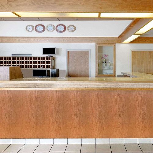 This image shows a hotel reception desk with wooden panels, a key holder rack, multiple clocks on the wall, and a tile floor.