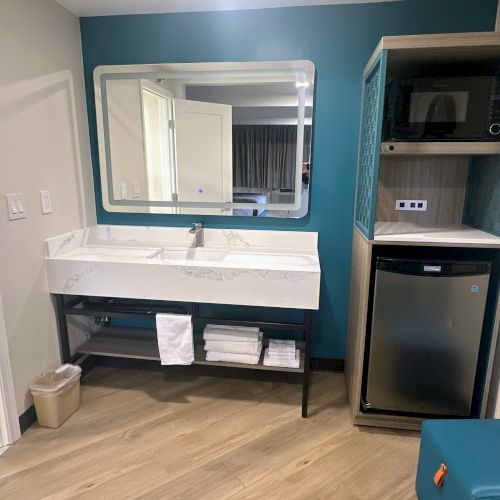 A hotel room setup with a sink, mirror, mini-fridge, microwave, and towels on a rack below the sink.