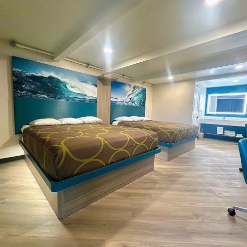 The image shows a modern hotel room with two large beds, wave artwork, a desk, and a bright mirror in the bathroom area.