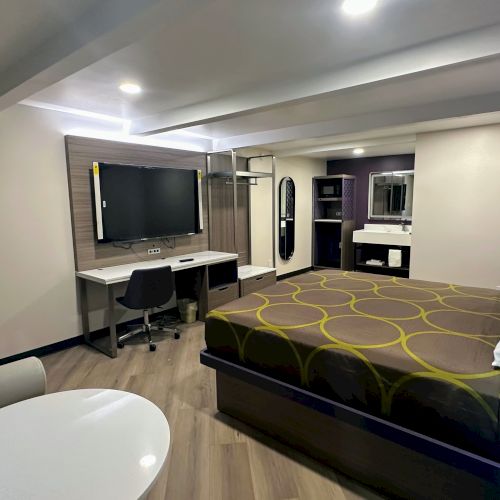 A modern hotel room with a round table, chair, TV, bed, and a bathroom vanity in a clean, minimalist design style.