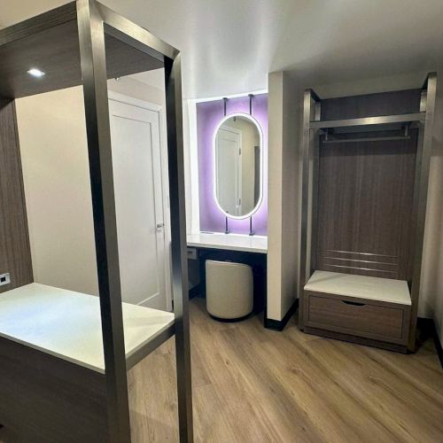 A modern room with a vanity, mirror with purple lighting, a wooden closet, and a desk with outlets. Minimalistic and functional design.