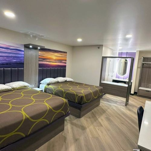 The image shows a modern hotel room with two beds, a wall-mounted mirror, desk, and a wardrobe, featuring a sunset beach mural.