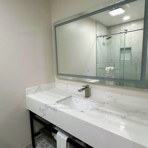 A modern bathroom with a large mirror, white vanity, under-sink shelves, and a shower with a glass door and marble design.