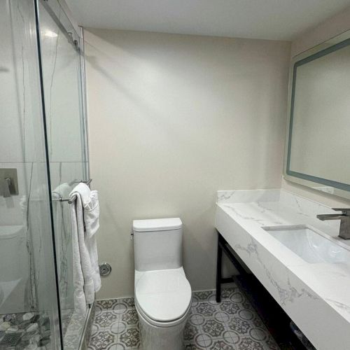 A modern bathroom featuring a toilet, patterned floor tiles, a glass shower, and a marble countertop sink area with a mirror.
