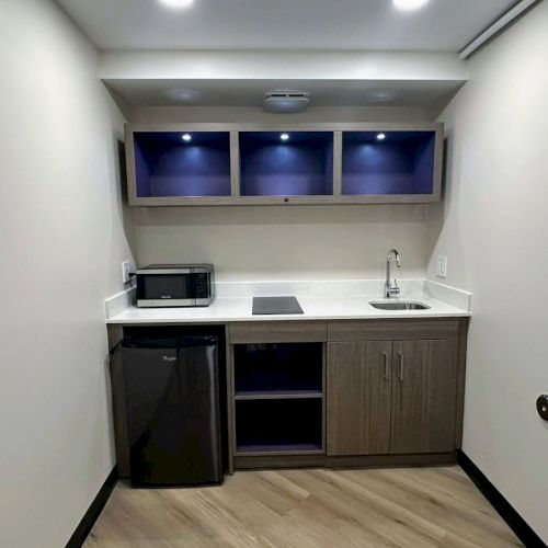A compact kitchenette with a fridge, microwave, sink, and storage cabinets, illuminated by overhead lights.