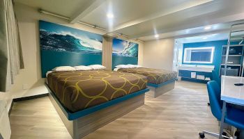 A modern hotel room with two beds, wave-themed artwork, a desk with a chair, and an illuminated mirror in the bathroom area.