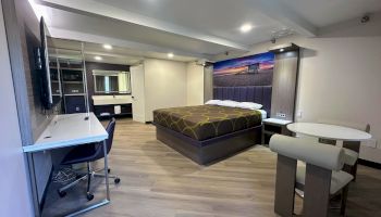 The image shows a modern hotel room with a bed, desk, TV, table, and chairs, featuring sleek decor and a beach scene artwork.