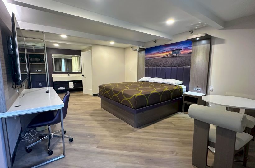 The image shows a modern hotel room with a bed, desk, TV, table, and chairs, featuring sleek decor and a beach scene artwork.