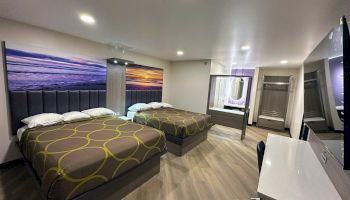 A modern hotel room with two double beds, sunset artwork, a desk, a mirror, and open wardrobe space, under soft overhead lighting.