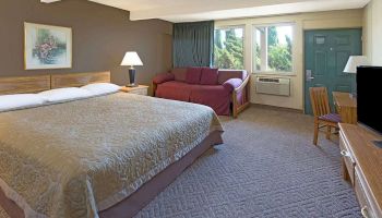 This image shows a hotel room with a king-sized bed, a red couch, a small table with chairs, a TV, a framed picture, and a door leading outside.