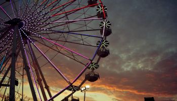 A Ferris wheel is illuminated against a colorful, cloudy sunset sky with scattered lights and structures in the foreground.