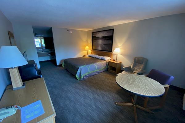 A hotel room with a king-size bed, desk, chairs, table, TV, lamps, and framed art on the wall. The room appears tidy and well-lit.