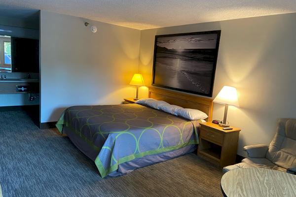 A hotel room with a king-sized bed, nightstands with lamps, a picture above the bed, a chair, a small round table, and a sink area.