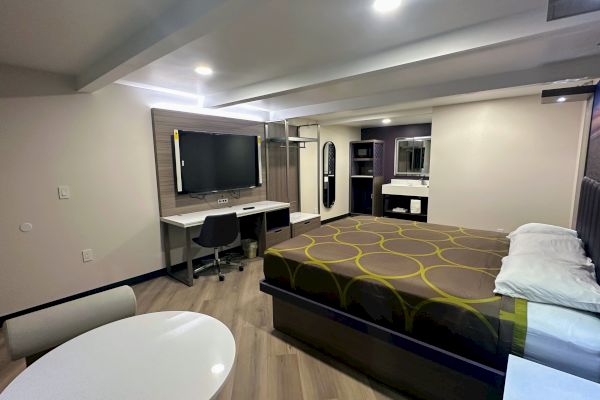 The image shows a modern hotel room with a bed, flat-screen TV, desk, and a table with chairs. A sink and mirror are in the corner.